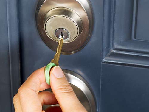 A close-up shows a hand inserting a brass key with a green rubber grip into the keyhole of a silver deadbolt lock set against a dark blue door.