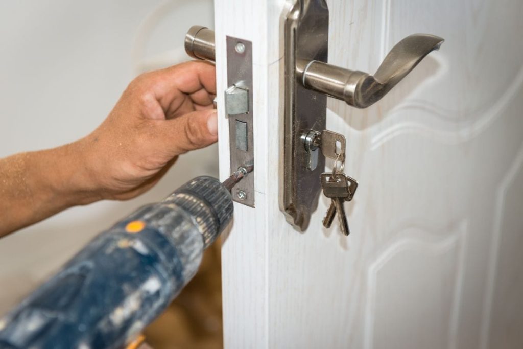 A close-up of a person's hands using a power drill to install a silver-colored door lock on a white door.