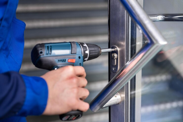 A man in blue overalls operates a drill on a door at a storefront specializing in lock repairs