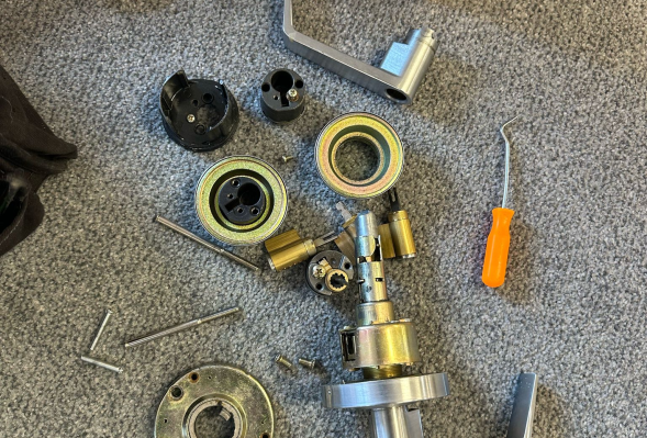 Disassembled door lock mechanism with various metal parts, screws, and a small orange-handled tool lying on a gray carpet.